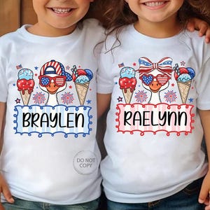 May include: White t-shirts with patriotic designs. One shirt features a goose wearing sunglasses and a hat, with the name "BRAYLEN" below. The other shirt has a goose with a bow and heart-shaped sunglasses, with the name "RAELYNN". Both shirts have ice cream cones and fireworks.
