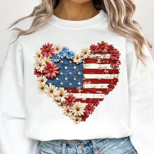 May include: White sweatshirt featuring a heart-shaped design of the American flag with a distressed wood effect. The flag is surrounded by red, white, and blue flowers. A patriotic themed apparel item.