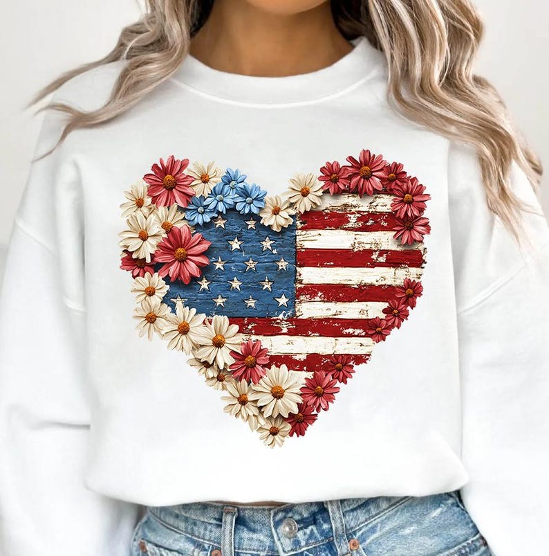 May include: White sweatshirt featuring a heart-shaped design of the American flag with a distressed wood effect. The flag is surrounded by red, white, and blue flowers. A patriotic themed apparel item.