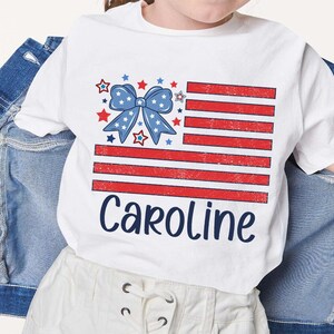 May include: White t-shirt with a red, white, and blue American flag design and a blue bow with stars. The text "Caroline" is printed below the flag.