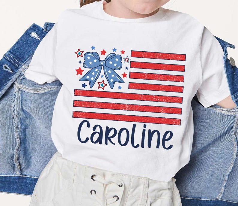 May include: White t-shirt with a red, white, and blue American flag design and a blue bow with stars. The text "Caroline" is printed below the flag.
