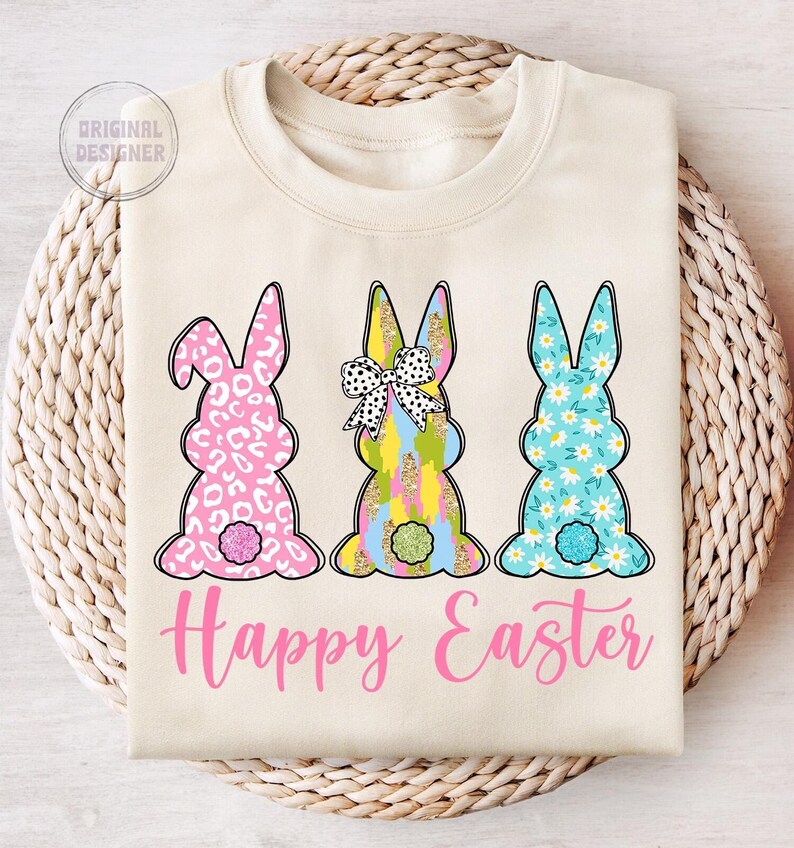 May include: A beige sweatshirt with three bunny silhouettes. The bunnies are decorated with different patterns, including leopard print, floral, and geometric designs. The bunnies are arranged in a row, and the text "Happy Easter" is printed below them in pink.