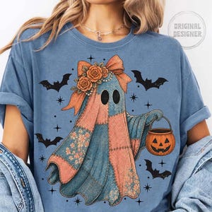 May include: A blue t-shirt featuring a patchwork ghost design with a bow, roses, and a pumpkin bucket. Black bats and stars are scattered around the ghost. The shirt has the words "Original Designer" in a circle.