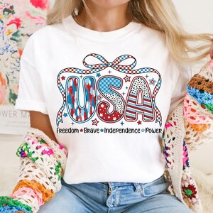 Peut inclure: T-shirt blanc avec un graphique USA rouge, blanc et bleu avec un n&oelig;ud et des &eacute;toiles. Le texte sous le graphique indique "Freedom Brave Independence Power".