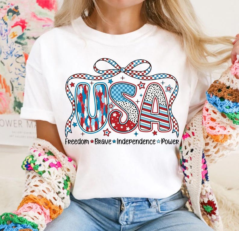 Peut inclure: T-shirt blanc avec un graphique USA rouge, blanc et bleu avec un n&oelig;ud et des &eacute;toiles. Le texte sous le graphique indique "Freedom Brave Independence Power".