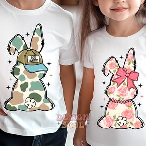 May include: Two white t-shirts with a bunny rabbit design. One bunny is camouflage with a blue and white baseball cap that says "Egg Hunter". The other bunny is pink and white with a pink bow.