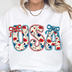May include: White sweatshirt with a red, white, and blue floral design that spells out "USA".