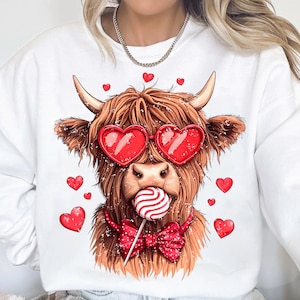 May include: White sweatshirt with a brown and white Highland cow wearing red heart-shaped sunglasses and a red bow tie. The cow is holding a red and white lollipop in its mouth. There are red hearts scattered around the cow.