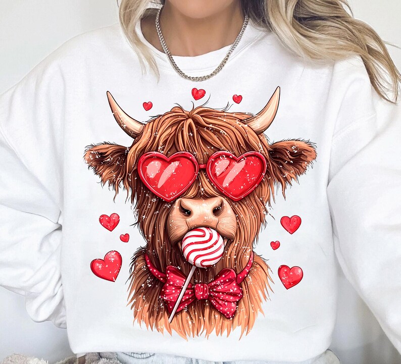 May include: White sweatshirt with a brown and white Highland cow wearing red heart-shaped sunglasses and a red bow tie. The cow is holding a red and white lollipop in its mouth. There are red hearts scattered around the cow.