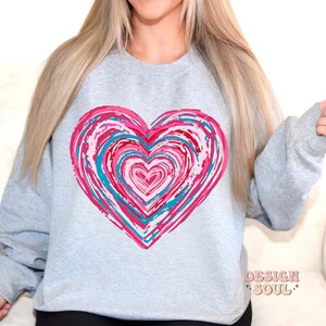 Pu&ograve; includere: Una felpa grigia con un grande disegno a forma di cuore rosa e blu. Il cuore &egrave; composto da cerchi concentrici di vernice rosa, blu e bianca.