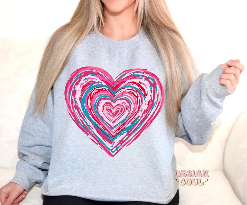 Pu&ograve; includere: Una felpa grigia con un grande disegno a forma di cuore rosa e blu. Il cuore &egrave; composto da cerchi concentrici di vernice rosa, blu e bianca.