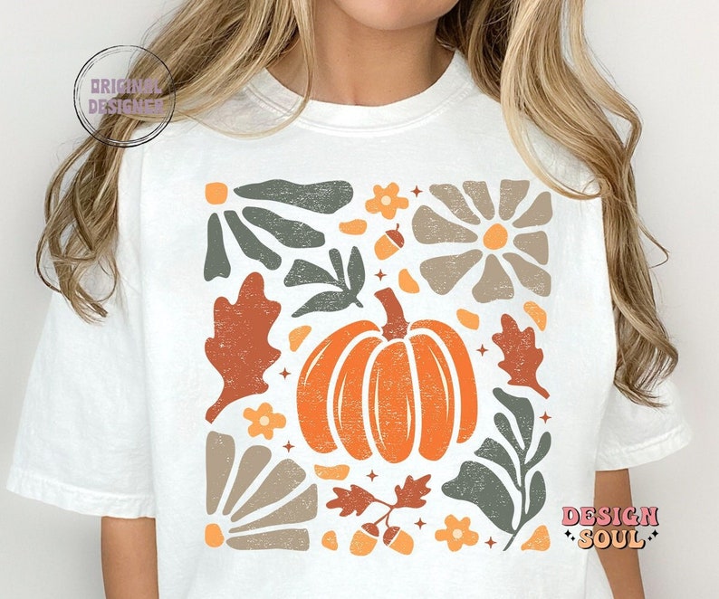 May include: White t-shirt featuring a graphic design with a pumpkin, leaves, acorns, and flowers in a retro style. The design is in shades of orange, brown, green, and beige.