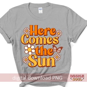 May include: A gray t-shirt with the text "Here Comes the Sun" in orange and yellow letters with a butterfly and daisies. The text is in a retro font style.