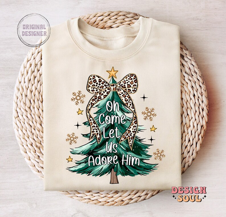 May include: A beige sweatshirt with a Christmas tree design. The tree is green with a leopard print bow and gold glitter accents. The text "Oh Come Let Us Adore Him" is written on the tree.