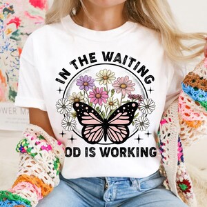 May include: White t-shirt with a graphic design of a butterfly and flowers in a circle. The text "In the waiting God is working" is printed below the circle.