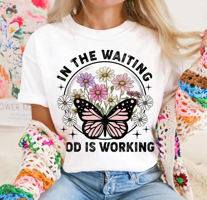 May include: White t-shirt with a graphic design of a butterfly and flowers in a circle. The text "In the waiting God is working" is printed below the circle.