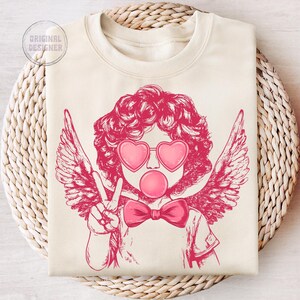 May include: A pink and white graphic design of a cherub with wings, wearing heart-shaped sunglasses and blowing a bubble. The cherub is giving a peace sign with one hand and has a pink bow tie.