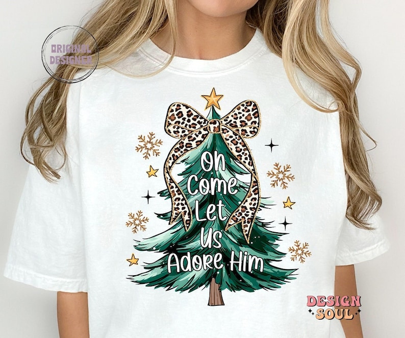 May include: White t-shirt with a green Christmas tree design. The tree is decorated with a leopard print bow and the words "Oh Come Let Us Adore Him".