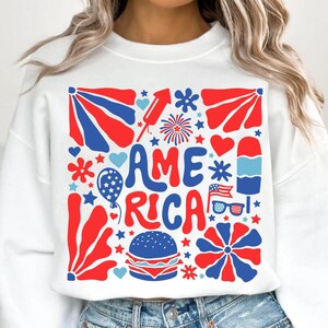 以下が含まれることがあります： 星、花火、ハート、その他の愛国的なイメージに囲まれた「AMERICA」という言葉が描かれた、赤、白、青のグラフィックデザインの白いスウェットシャツです。