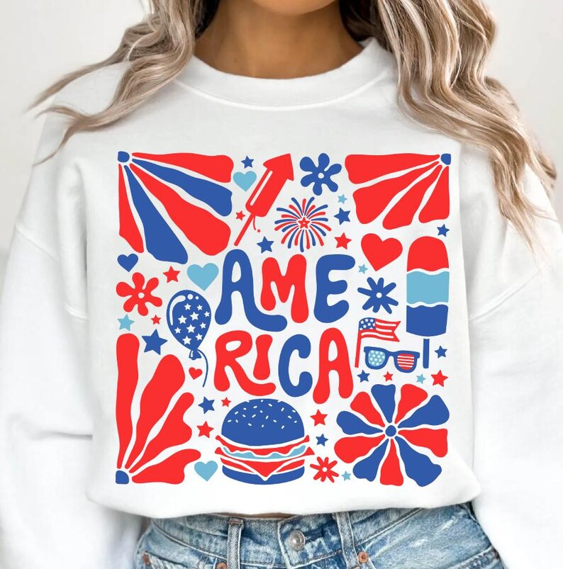 以下が含まれることがあります： 星、花火、ハート、その他の愛国的なイメージに囲まれた「AMERICA」という言葉が描かれた、赤、白、青のグラフィックデザインの白いスウェットシャツです。