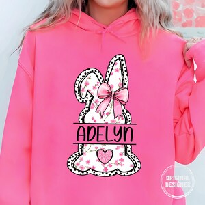 Pu&ograve; includere: Felpa rosa con un grafico di coniglio bianco con fiori rosa e un fiocco rosa. Il grafico del coniglio ha pois neri e il testo "Adelyn" &egrave; scritto sotto il coniglio.