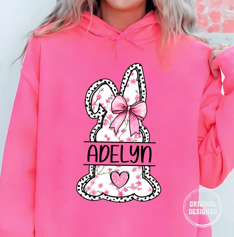 Pu&ograve; includere: Felpa rosa con un grafico di coniglio bianco con fiori rosa e un fiocco rosa. Il grafico del coniglio ha pois neri e il testo "Adelyn" &egrave; scritto sotto il coniglio.