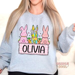 May include: A light gray sweatshirt with a design of three Easter bunnies sitting on a pink and white checkered background. The bunnies are wearing colorful clothes and have a pink, white, and green color scheme. The text "Olivia" is written in pink on the checkered background.