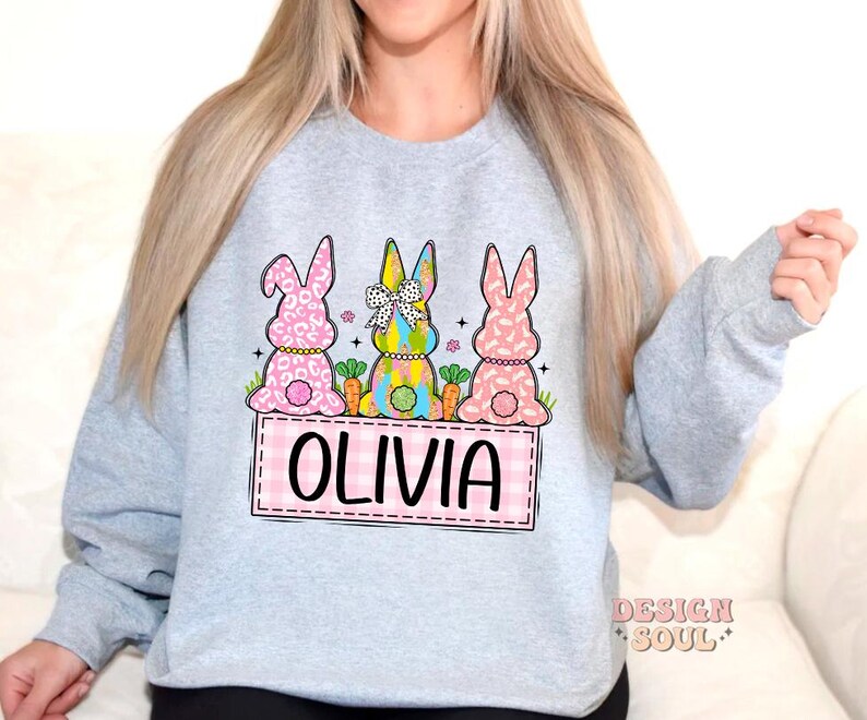 May include: A light gray sweatshirt with a design of three Easter bunnies sitting on a pink and white checkered background. The bunnies are wearing colorful clothes and have a pink, white, and green color scheme. The text "Olivia" is written in pink on the checkered background.