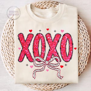 May include: A cream-colored sweatshirt with a pink sequined "XOXO" design and a black and white polka dot bow. The sweatshirt has a crew neck and long sleeves.