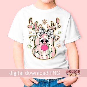 May include: A white t-shirt with a pink and green reindeer design. The reindeer has a black and white polka dot bow. The design includes gold glitter accents and snowflakes.