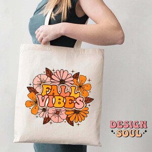 May include: A white tote bag featuring a floral design and the text "Fall Vibes" in a retro font. The flowers are orange, pink, and yellow. The bag is being held by a person.