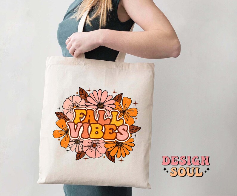 May include: A white tote bag featuring a floral design and the text "Fall Vibes" in a retro font. The flowers are orange, pink, and yellow. The bag is being held by a person.