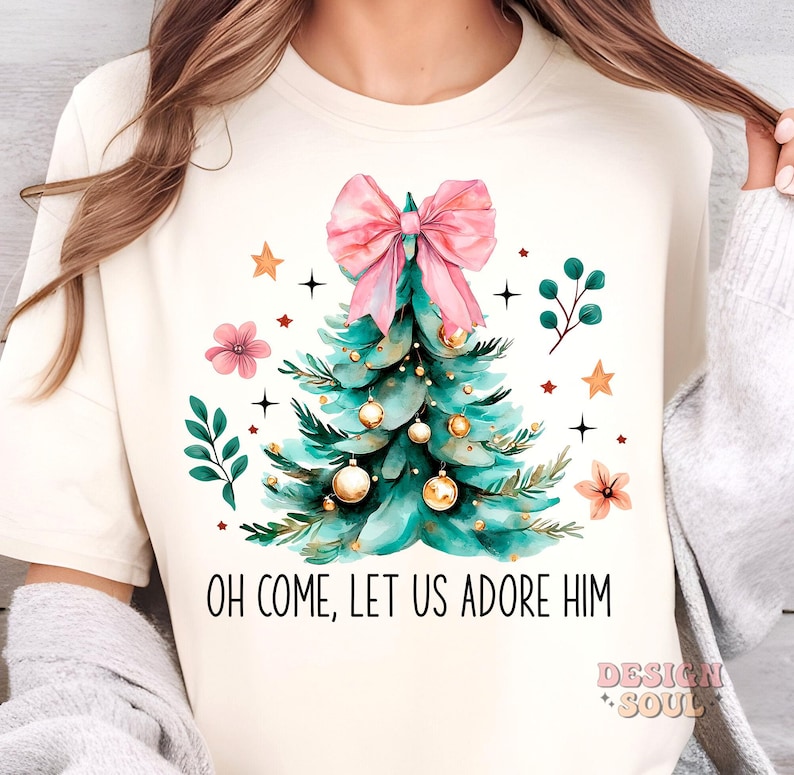 May include: A white t-shirt with a watercolor illustration of a Christmas tree with a pink bow, gold ornaments, and green leaves. The text "Oh Come, Let Us Adore Him" is printed below the tree.