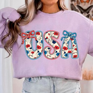 May include: A lavender sweatshirt with a floral pattern "USA" graphic. The letters are outlined in red, white, and blue.