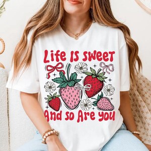 May include: White t-shirt with a graphic print of strawberries and flowers. The text "Life is sweet and so are you" is printed on the shirt.