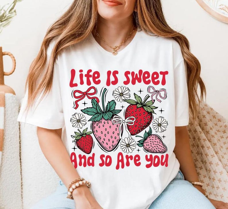 May include: White t-shirt with a graphic print of strawberries and flowers. The text "Life is sweet and so are you" is printed on the shirt.