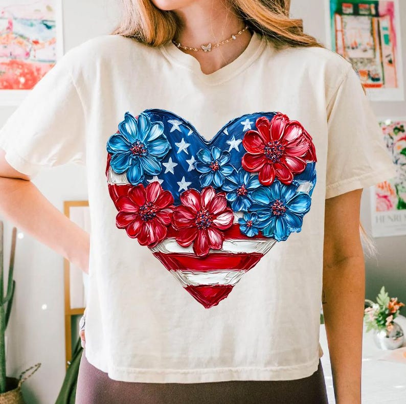 May include: A white t-shirt with a red, white, and blue heart-shaped design. The heart is made up of flowers and the American flag.