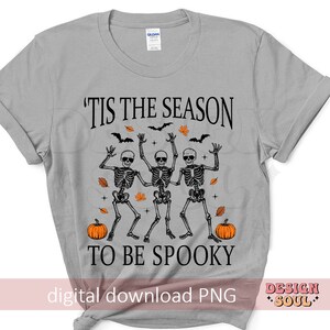 May include: A grey t-shirt with the text "'Tis the season to be spooky" in black. Three skeletons are dancing with pumpkins on either side of the text.