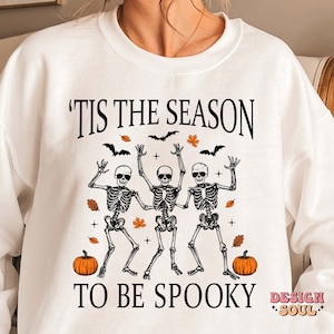 May include: White sweatshirt with the text 'Tis the season to be spooky' and a graphic of three skeletons dancing with pumpkins and autumn leaves.