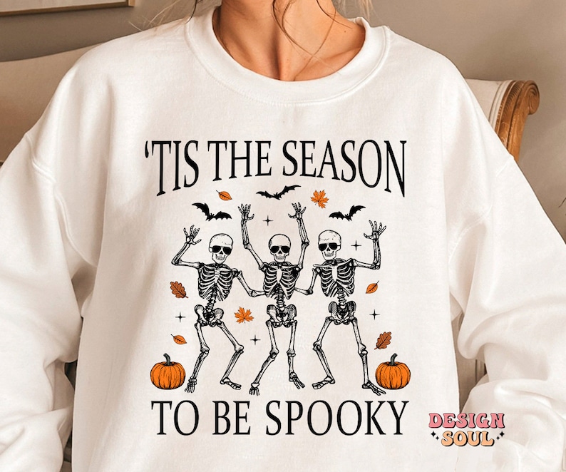 May include: White sweatshirt with the text 'Tis the season to be spooky' and a graphic of three skeletons dancing with pumpkins and autumn leaves.
