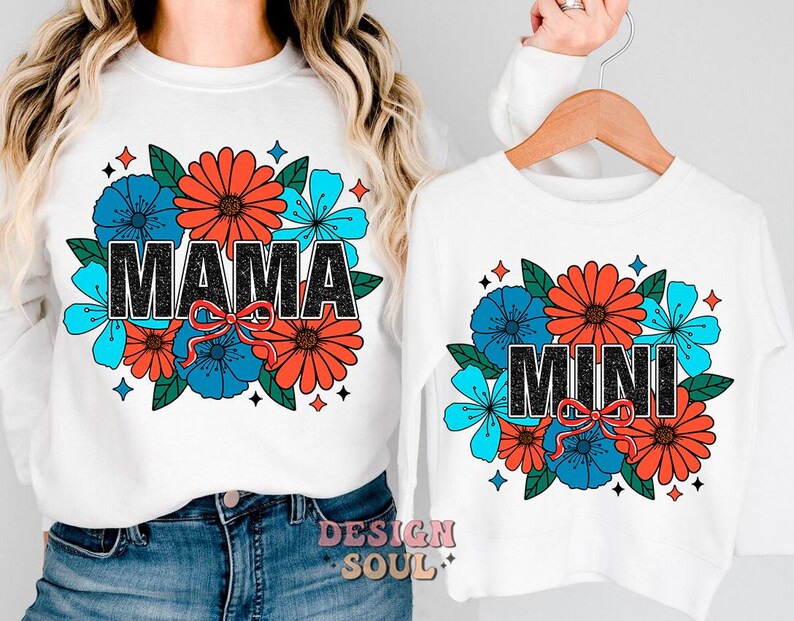 Pu&ograve; includere: Due magliette bianche con un design floreale e il testo "Mama" e "Mini" in nero. I fiori sono rossi, blu e arancioni.