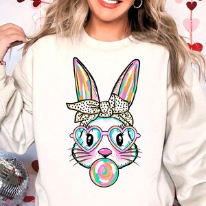 May include: A white sweatshirt featuring a colourful cartoon bunny wearing heart-shaped sunglasses and a polka dot headband. The bunny is holding a colourful lollipop in its mouth.