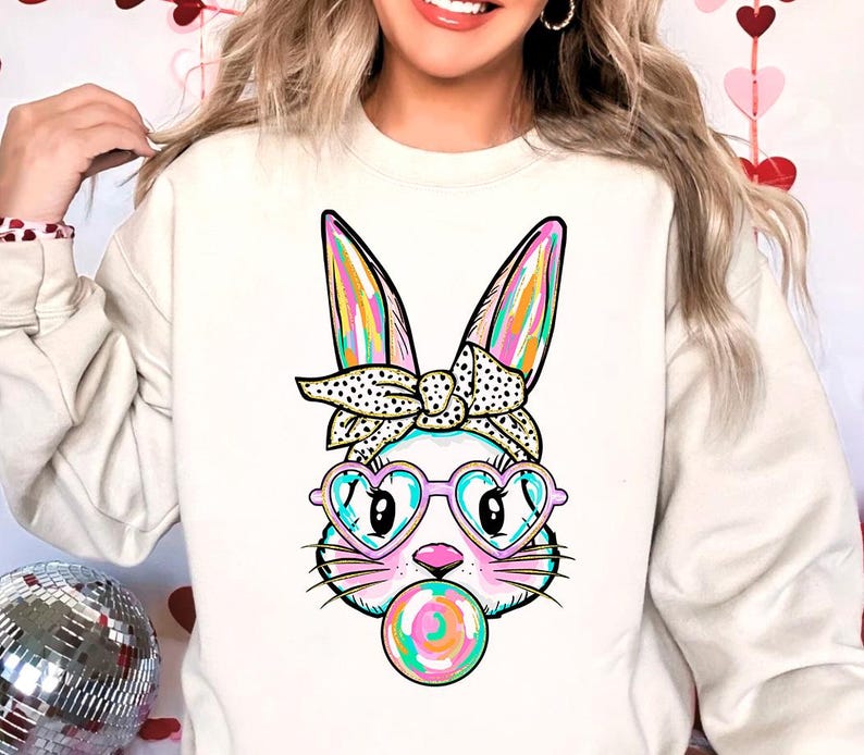 May include: A white sweatshirt featuring a colourful cartoon bunny wearing heart-shaped sunglasses and a polka dot headband. The bunny is holding a colourful lollipop in its mouth.