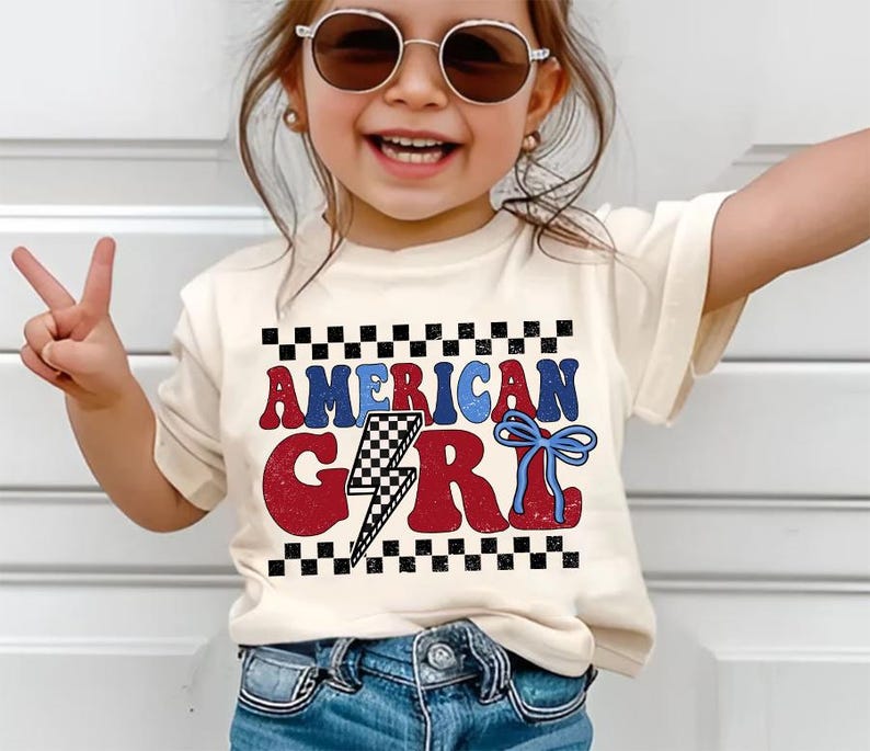 May include: A white t-shirt with a red, white, and blue graphic that says "AMERICAN GIRL" with a lightning bolt and a blue bow. The shirt is being worn by a child wearing blue jeans and sunglasses.
