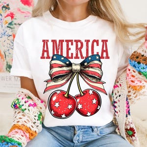May include: White t-shirt with a red, white, and blue bow with cherries and the word "AMERICA" printed on it.