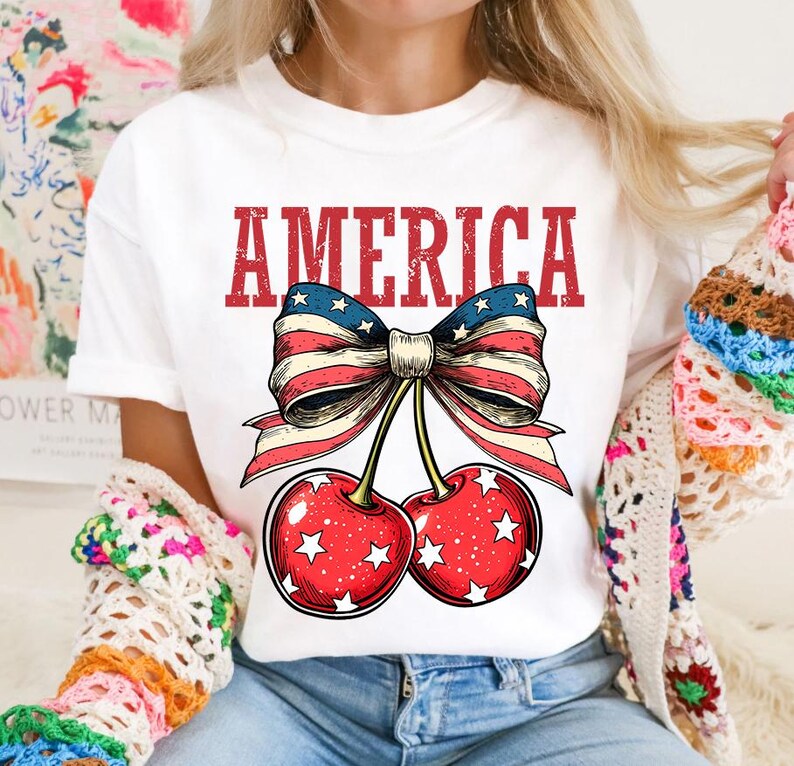 May include: White t-shirt with a red, white, and blue bow with cherries and the word "AMERICA" printed on it.