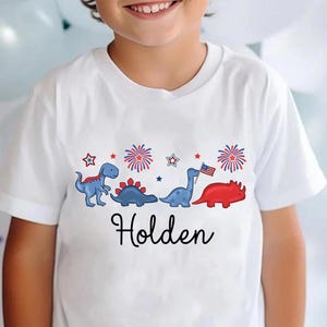 May include: White t-shirt featuring a patriotic dinosaur design with the name "Holden" in black script. The design includes blue and red dinosaurs, fireworks, and stars, with one dinosaur holding an American flag.