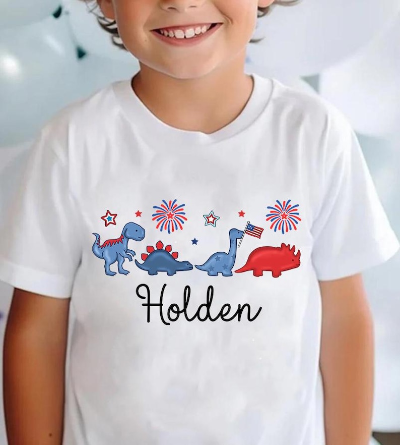 May include: White t-shirt featuring a patriotic dinosaur design with the name "Holden" in black script. The design includes blue and red dinosaurs, fireworks, and stars, with one dinosaur holding an American flag.