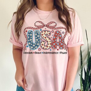 May include: Pink t-shirt with a red, white, and blue USA graphic with a bow and stars. The text below the graphic reads "Freedom * Brave * Independence * Power".