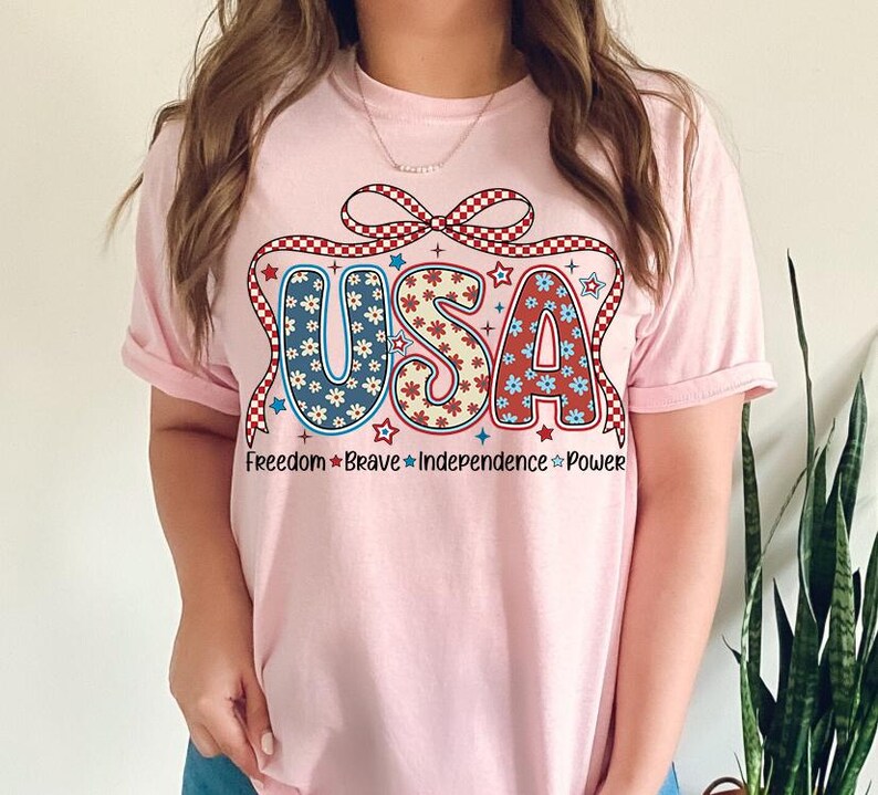 May include: Pink t-shirt with a red, white, and blue USA graphic with a bow and stars. The text below the graphic reads "Freedom * Brave * Independence * Power".